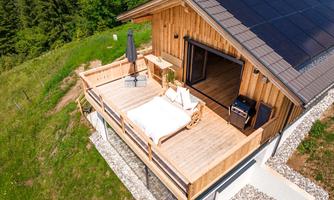 A modern wooden cabin with a large terrace. The terrace is equipped with a lounge chair and a sun umbrella, surrounded by green meadows.