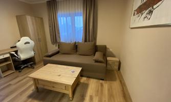 A cozy living space with a brown sofa and a wooden table. A picture hangs on the wall, and next to the sofa is a desk chair.