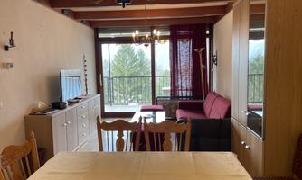 A cozy living room with a dining area and a television. Large windows offer a view of the surroundings.