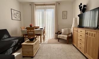 A cozy living room with a gray sofa, a wooden table, and chairs. Large windows let in a lot of light, and there is a television as well as a dresser.