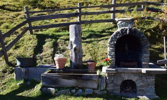 A rustic stone oven with potted plants and a simple water basin. In the background, a wooden railing and a green meadow are visible.