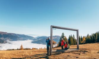 A group of people is posing in front of a large picture frame in the mountains. In the background, gentle hills and mist can be seen.