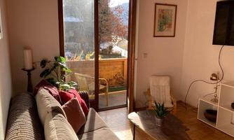 A cozy living room with a sofa, a table, and plants. Windows overlooking a balcony and a sunny view.