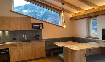 A modern kitchen corner with wooden paneling and large windows that offer a view of the mountains. The dining area is designed to be cozy and inviting.
