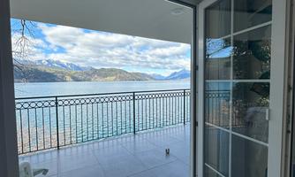 An open window with a view of a lake and the surrounding mountains. The terrace is bright and welcoming, with a chair and a rug.