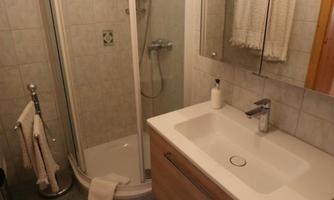 A small bathroom with a shower and a modern sink. The walls are tiled and there is a towel rack as well as a bath mat.