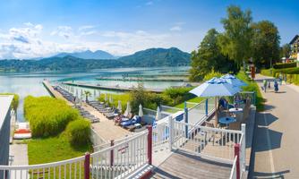 A picturesque lake with a view of rolling hills and blue sky. At the shore, there are loungers and umbrellas, surrounded by green vegetation.