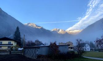 A picturesque mountain landscape with partially fog-covered hills. In the background, snow-capped peaks and a clear sky can be seen.