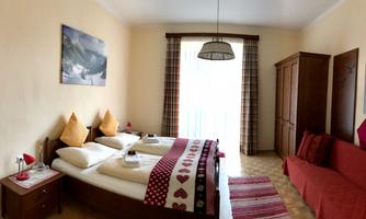 A cozy room with a double bed, a sofa, and a large window. The warm color scheme and decorative cushions create an inviting atmosphere.