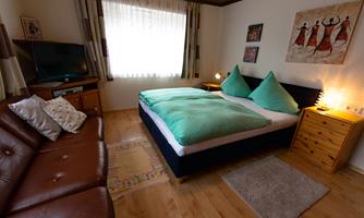 A cozy bedroom with a double bed and large, green pillows. Next to the bed is a TV and a wall decoration featuring dancers.