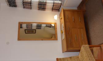 A bright room with a wooden wardrobe and a large mirror on the wall. Next to the wardrobe is a chair made of woven material.