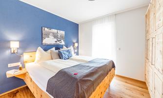 A cozy bedroom with a large bed and blue walls. Daylight streams through the window, creating a bright atmosphere.