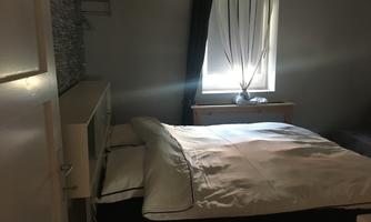 A cozy bedroom with a large bed and a window that lets in daylight. The walls are painted in soft colors and there is a small shelf.