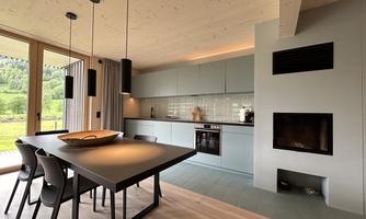 A modern kitchen with a large dining table and black chairs. Large windows offer a view of the green landscape.