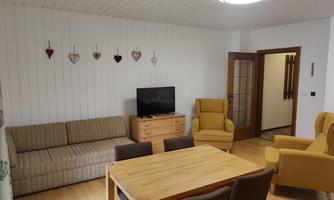 A cozy living room with a sofa, two yellow chairs, and a dining table. Decorative hearts hang on the wall, and a television stands on a wooden cabinet.