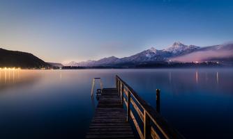 A tranquil lake with a wooden pier and majestic mountains in the background. The sky is tinted in gentle colors at dusk.