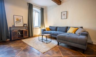 A cozy living room with a gray sofa and decorative cushions. The room has large windows and a beautiful wooden floor.