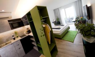 A modern room with an open kitchen and a large window. Green accents and bright furniture create a friendly atmosphere.