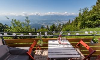A cozy terrace with red chairs and a table, surrounded by green trees. In the background, a stunning mountain landscape stretches out under a clear sky.