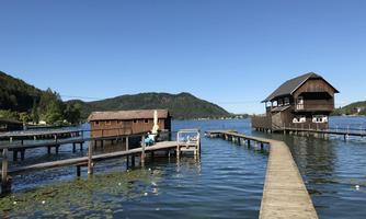 A tranquil lake with wooden piers and two charming boathouses. In the background, gentle hills and a blue sky can be seen.