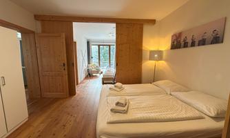 A bright bedroom with a double bed and a bedside table. In the background, there is a window overlooking a cozy seating area.