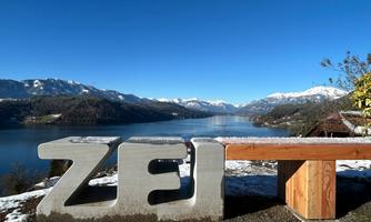A large, graphic seating area with the letters "ZEI" stands in front of a picturesque lake. In the background, snow-covered mountains are visible under a clear blue sky.