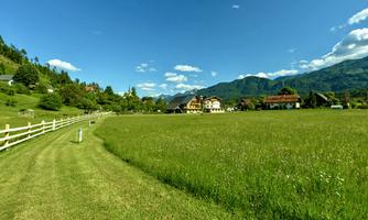 A spacious meadow surrounded by mountains and trees. In the background, some traditional houses can be seen.