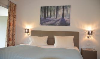A cozy bedroom with a large bed and two pillows. A picture of a forest with purple flowers hangs on the wall.