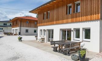 A modern building with wooden cladding and large windows. In front of the house is a terrace with tables and chairs.