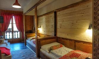 A cozy room with two wooden beds and warm light. The windows are decorated with red curtains and offer a view outside.