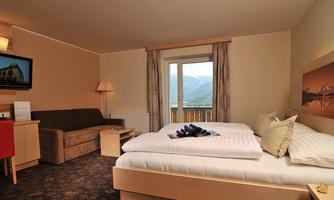 A comfortable hotel room with a double bed, a couch, and a table. Large windows offer a view of the mountains.