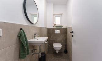 A modern bathroom with a white sink and a round mirror. The walls are tiled in neutral colors, and a green towel hangs next to the sink.