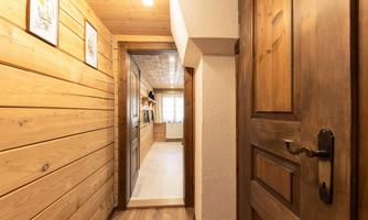 A cozy hallway with wooden walls and a wooden door. At the end of the hallway, a bright room with windows is visible.