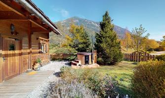A cozy wooden house in a rural setting with a view of the mountains. The garden is green and well-maintained, surrounded by trees.