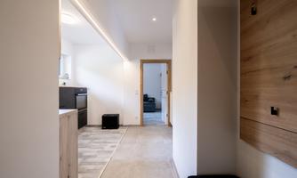 A modern hallway with a light floor and wooden shelf. In the background, a kitchen and a living area are visible.