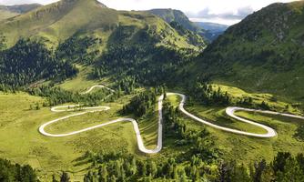A winding road meanders through a green mountain landscape. The surroundings are characterized by dense forests and gentle hills.