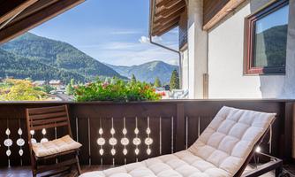A quiet balcony with a lounge chair and a view of the mountains. The sky is clear and the surroundings are green.
