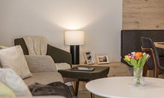 A cozy living room with comfortable sofas and a decorative lamp. On a table, there are flowers in a vase and some picture frames.