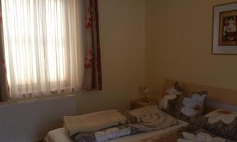 A cozy bedroom with a large window and light curtains. The bed is decorated with floral bedding and there is a nightstand with a lamp.
