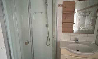 A modern bathroom with a clear shower cabin and a sink. The walls are tiled with white tiles, and a small mirror hangs above it.
