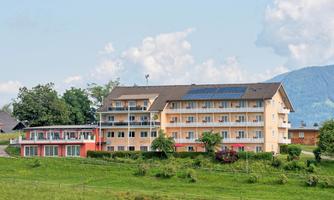 A modern building with several floors and large windows stands in a green landscape. The surroundings are characterized by mountains and a blue sky.