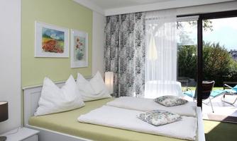 A bright bedroom with a double bed and two large pillows. The room has a balcony overlooking the pool area and is decorated with colorful pictures.