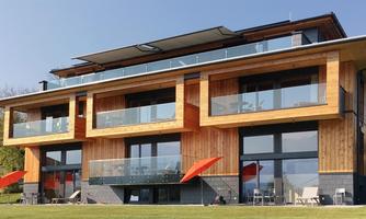A modern wooden house with large windows and balconies. In the foreground, there are green meadows and red sun umbrellas.