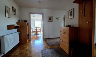 A bright hallway with wooden furniture and a carpet. In the background, there is a dining area with a table and chairs.