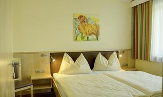 A bright hotel room with two single beds and a colorful painting on the wall. The room has large windows with curtains in warm colors.