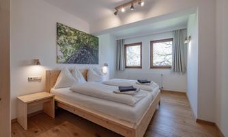 A modern bedroom with a double bed and two windows. The walls are painted light, and a large piece of artwork hangs above the bed.