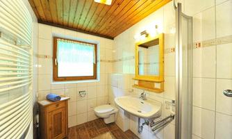 A modern bathroom with white tiles and a wooden ceiling. It is equipped with a sink, a shower, and a window.