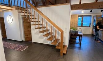 A modern and bright entrance with a wooden staircase. The room is open and simply designed, with tiled flooring and large windows.
