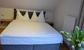 A cozy bedroom with a large bed and two pillows. Next to the bed is a small flower in a vase.