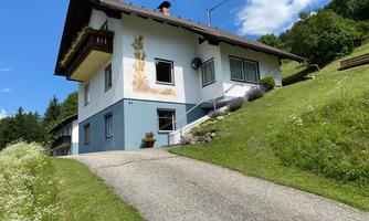 A beautiful house with a small slope and a well-maintained garden. Surrounded by trees and a clear, blue sky.